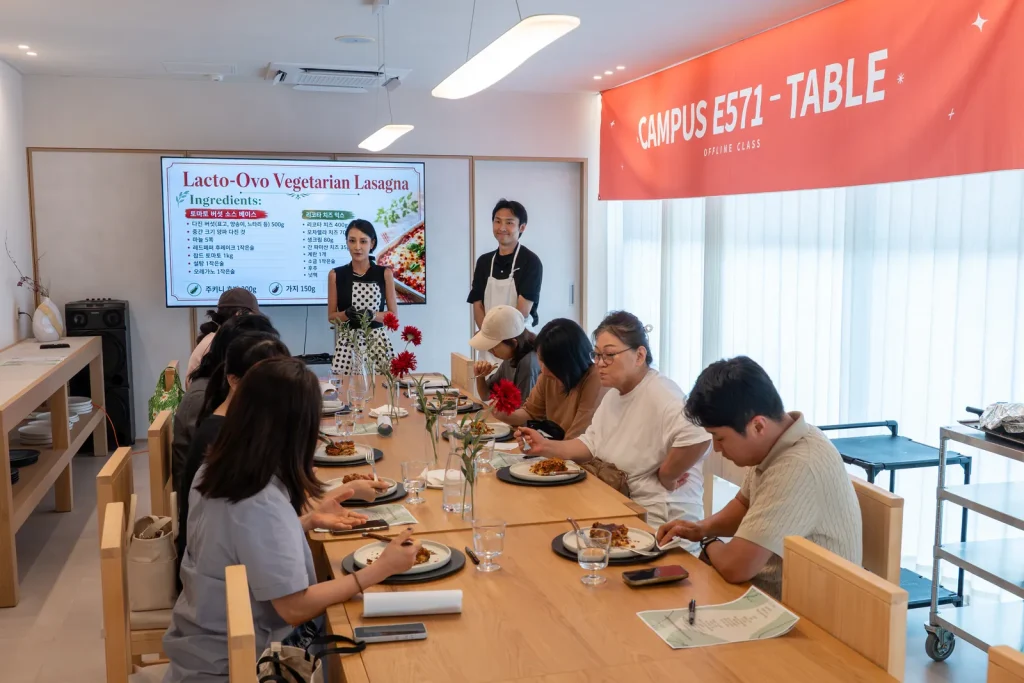 CAMPUS E571 – TABLE 현수막과 재료 목록을 띄운 스크린 앞, 접시가 놓인 긴 테이블.